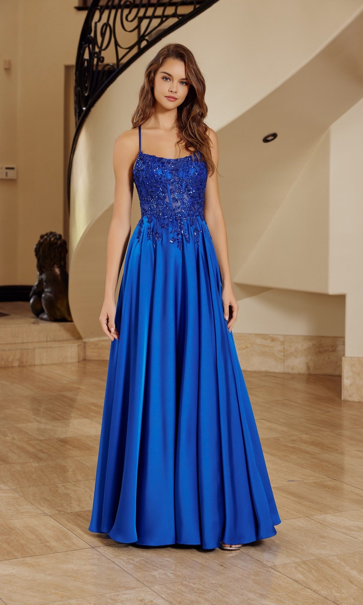 A woman is indoors wearing the Nox Anabel A1725 long prom dress in royal blue, featuring a sleeveless lace bodice and a flowing satin A-line skirt.